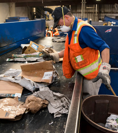 A worker in safety vest and mask sorts paper recycling